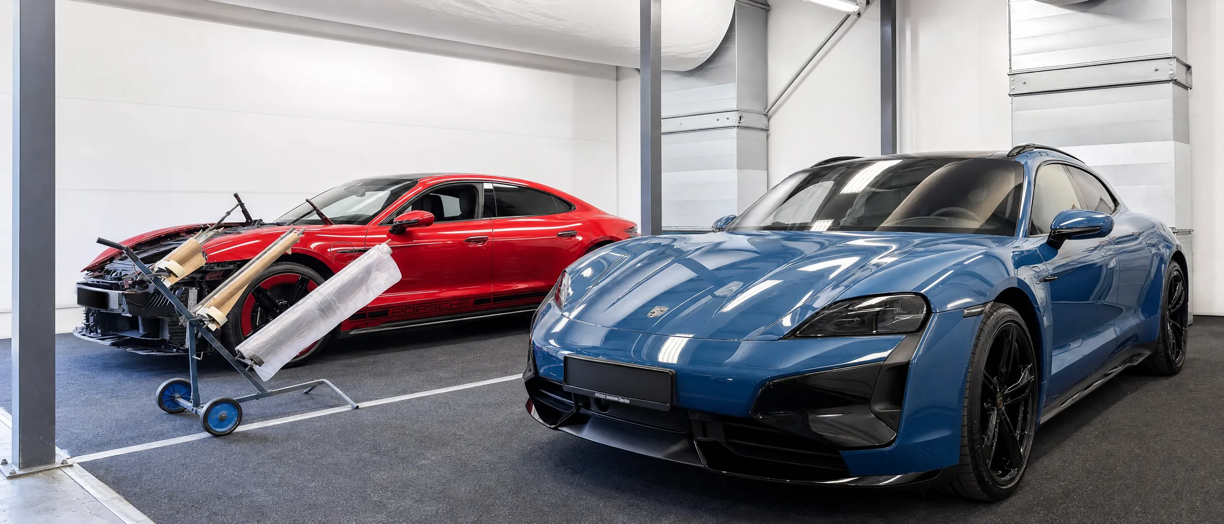 Zwei Porsche Taycan in der Werkstatt des CarCare Center Leipzig: ein blauer Wagen nach der Aufbereitung, dahinter ein roter Wagen mit demontierter Front in der Unfallinstandsetzung