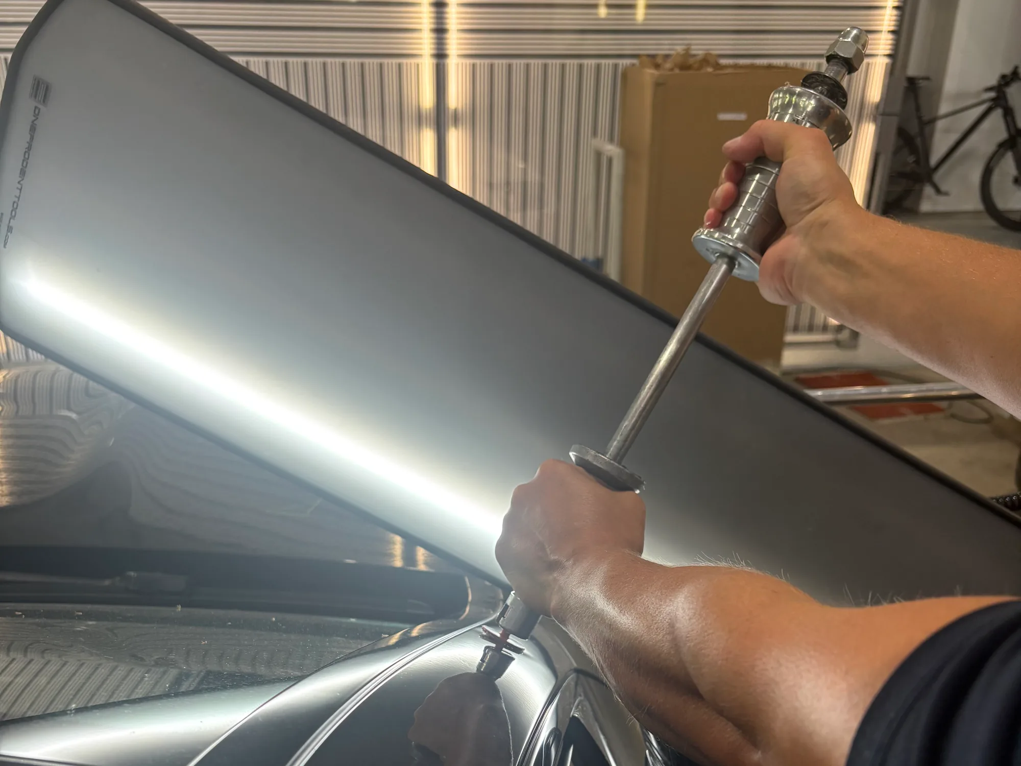 Techniker zieht unter dem Leuchtschirm mit Gleithammer und Klebepad eine Delle an der Dachsäule heraus, Hagelschadenreparatur im CarCare Center Leipzig